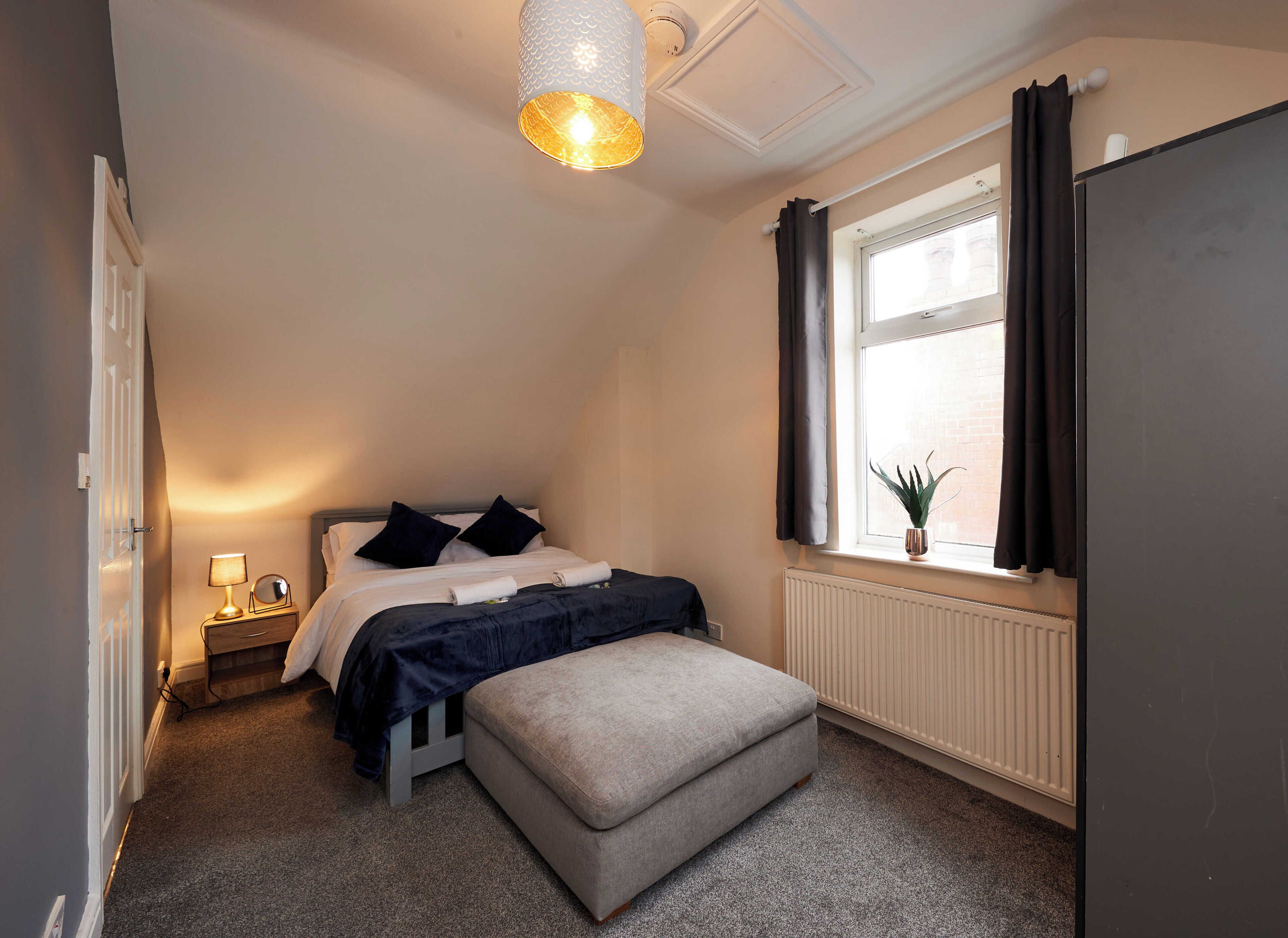 Une chambre à coucher comprenant un lit, une table de chevet, une lampe, une fenêtre avec des rideaux, un radiateur et une plante en pot.
