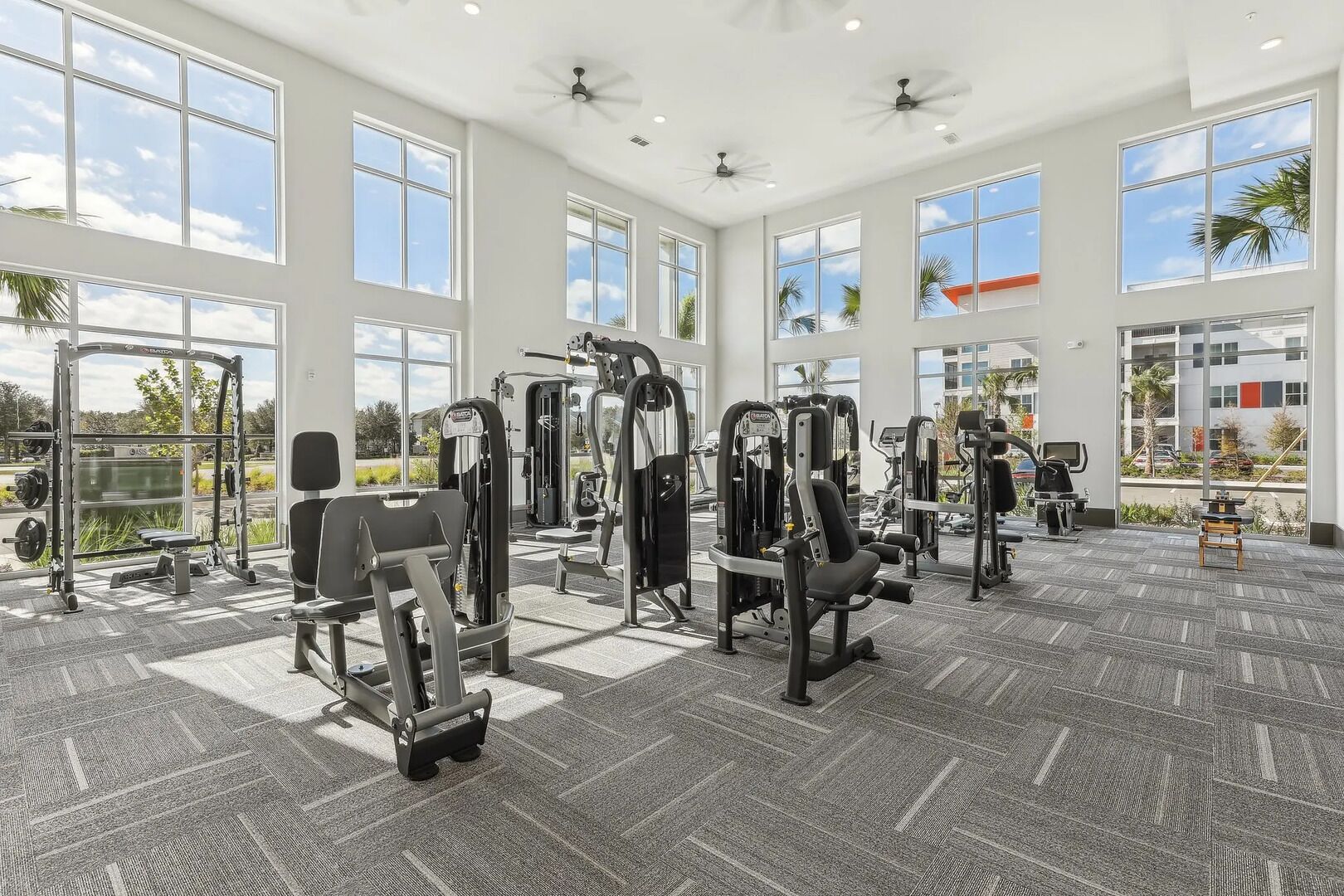 Une salle de sport spacieuse, équipée de divers appareils d’exercice, dotée de grandes fenêtres et d’un ventilateur au plafond.