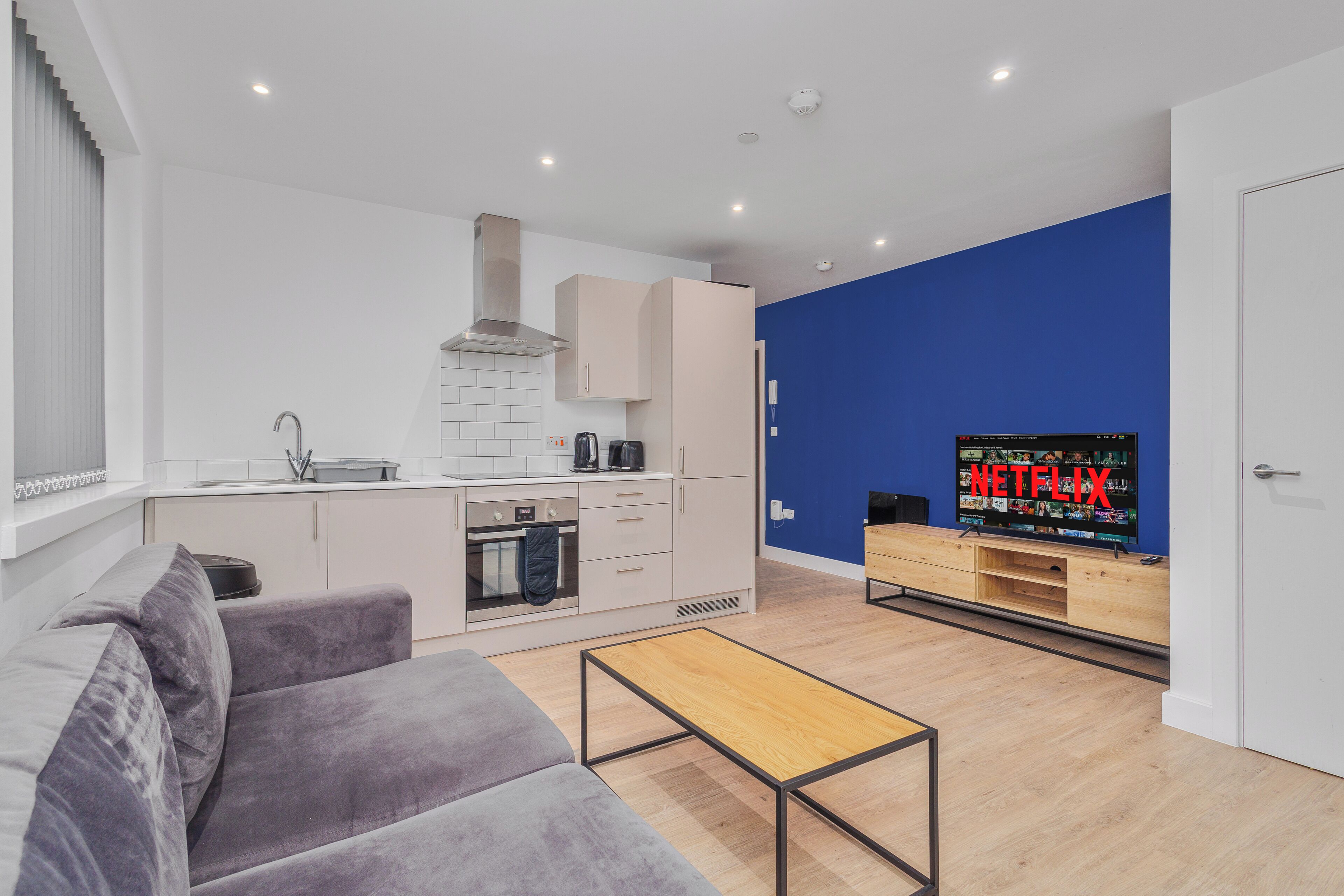 Een moderne woonkamer met een grijze bank, een houten salontafel, een keuken met witte kasten, een blauwe accentmuur en een televisie waarop 'NETFLIX' wordt weergegeven.