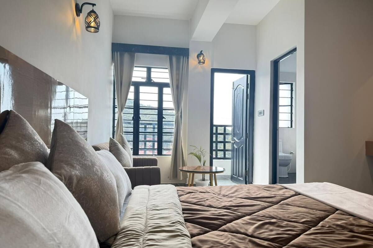 Un dormitorio con una cama grande, una mesita con una planta y vistas al exterior desde una puerta.