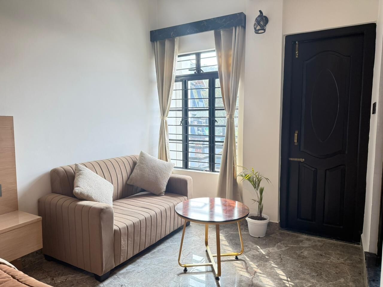 Una sala de estar con un sofá, una mesa redonda, una planta en maceta, una ventana con cortinas y una puerta negra.