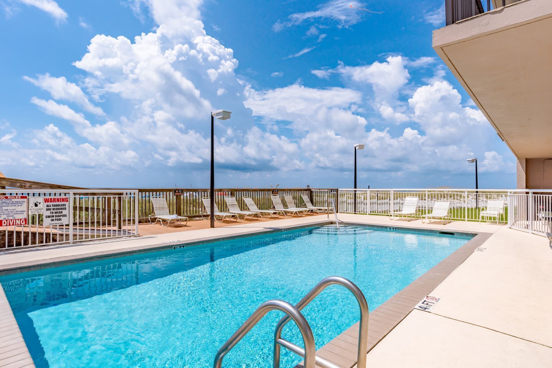 A swimming pool with a metal ladder, surrounded by lounge chairs and a fenced deck area.