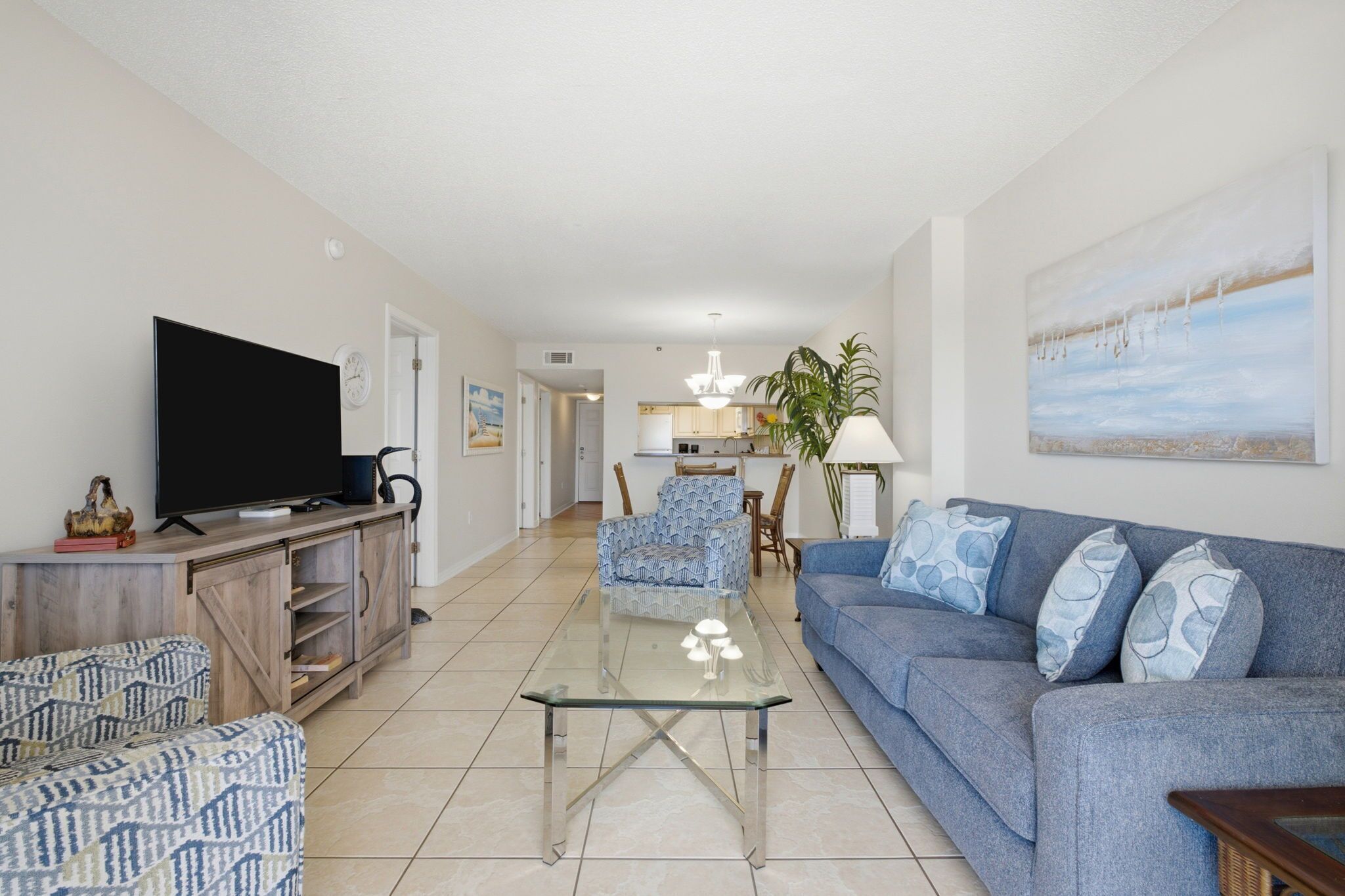 A living room with a TV, sofa, patterned chairs, a glass coffee table, and a kitchen in the background.