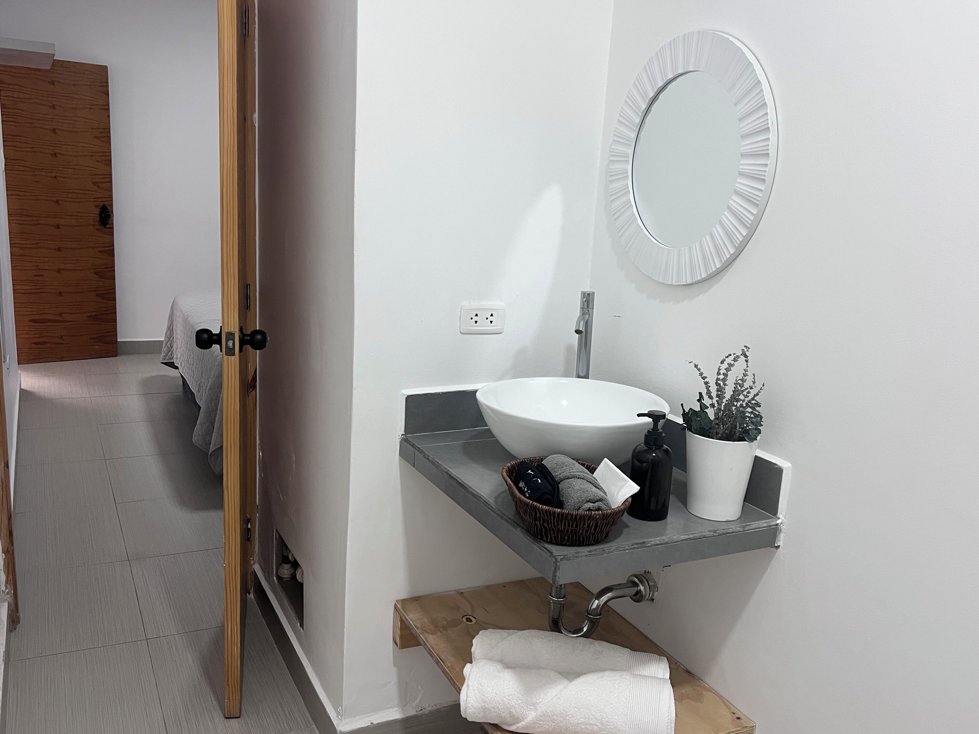 Ein Badezimmer mit einem weißen Waschbecken, einem runden Spiegel, einem Handtuchhalter und einer Topfpflanze.