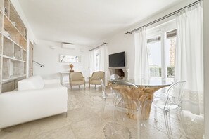 Ein modernes Wohnzimmer mit einem weißen Sofa, einem Glastisch und transparenten Stühlen. Ein Kamin und ein Flachbildfernseher sind an der Wand befestigt.