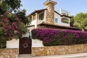 Ein zweistöckiges Haus mit einem Steinkamin, einem Balkon und einer Steinmauer mit violetten Blumen.