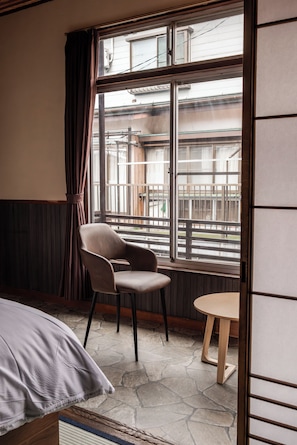 A room with a bed, a chair, a small table, and a large window with a view of buildings outside. - Snow Monkey Lodge - Hostel  (Yamanouchi)
