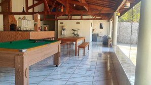 A pool table in a room with a tiled floor, a wooden bench, and a kitchen area with a refrigerator and cabinets. - Cozy country house surrounded by nature, with a beautiful sunset view. (Piracicaba)
