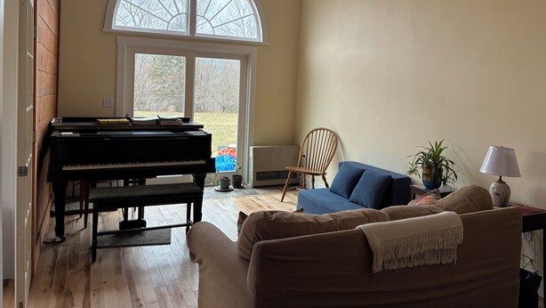 A living room with a piano, a blue sofa, a wooden chair, a lamp, and a view through a large window. - Charming 1-bedroom apartment in Worcester, private swimming hole. (Worcester)
