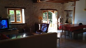 A rustic interior with a stone wall, wooden beams, a television, a sofa, a dining table, and a chair. - Charentais country house surrounded by vineyards with heated swimming pool (Mosnac)