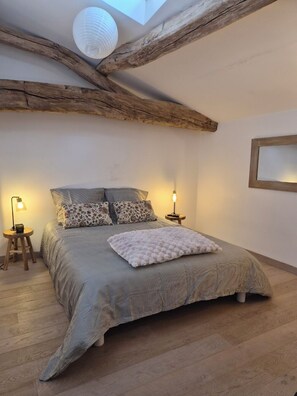 A bedroom with a sloped ceiling, exposed wooden beams, a bed with patterned bedding, two bedside lamps, and a round wall-mounted light fixture.
