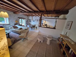 A cozy living room with a stone wall, wooden ceiling beams, a sofa, a dining table, and a bookshelf.