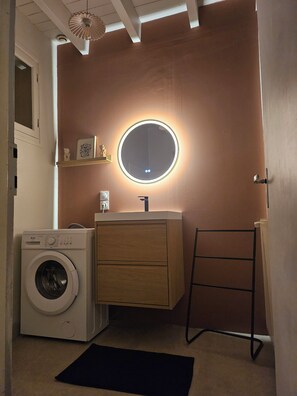A compact laundry room with a washing machine, a vanity with a mirror, and a black mat on the floor.