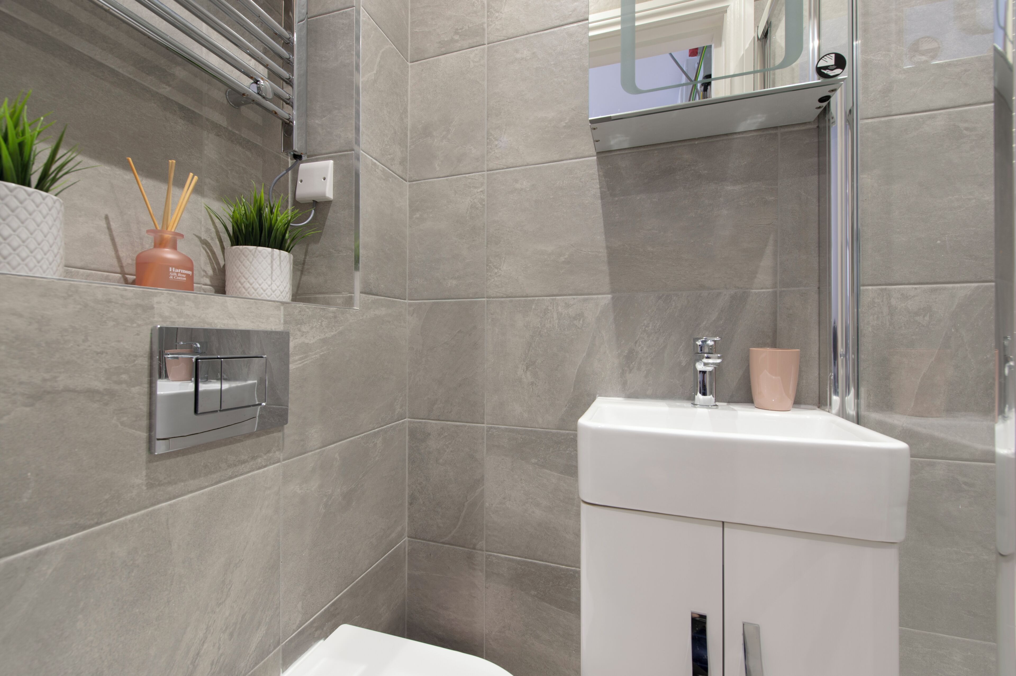 A modern bathroom with grey tiles, a white sink, a glass shower enclosure, and decorative plants.