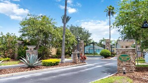 A gated entrance to a resort with a stone sign, palm trees, and a clear blue sky. - New Single-Level Private Pool Home with Game Room | Gated Community | 2 Master Bedrooms | Sleeps 10 (Davenport)