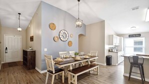 A dining area with a wooden table and chairs, a clock on the wall, and a kitchen in the background. - New Single-Level Private Pool Home with Game Room | Gated Community | 2 Master Bedrooms | Sleeps 10 (Davenport)