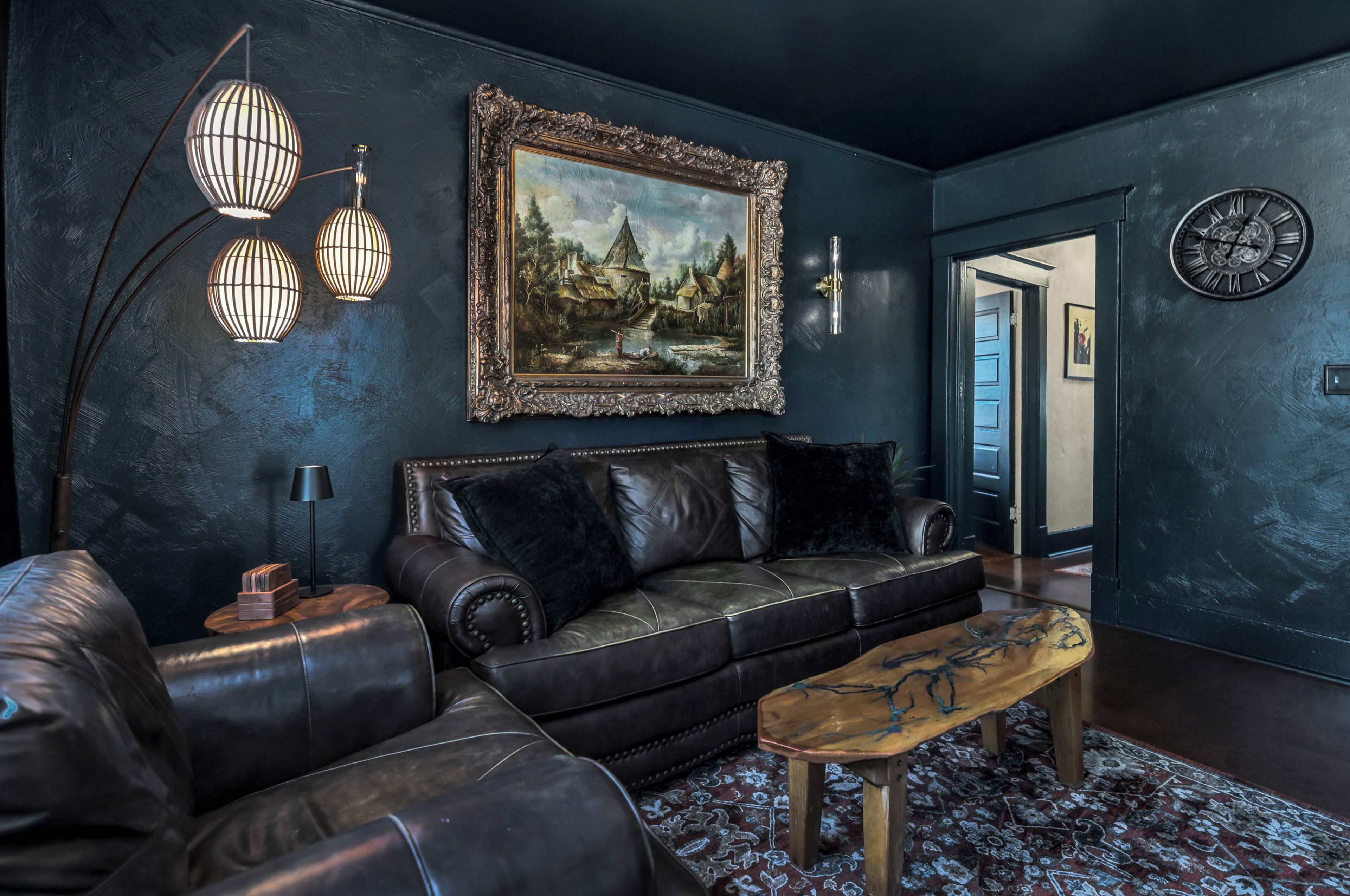 A dark-themed living room with a leather sofa, a wooden coffee table, a framed landscape painting, and a wall-mounted clock.