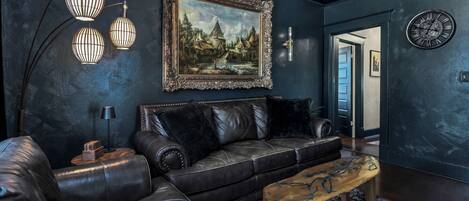A dark-themed living room with a leather sofa, a wooden coffee table, a framed landscape painting, and a wall-mounted clock.