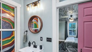 Ein Badezimmer mit einem weißen Waschbecken, einem runden Spiegel und einem farbenfrohen Fenster.