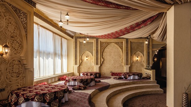 A room with ornate wall designs, a curved staircase, and tables with patterned tablecloths.