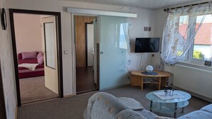 A room with a sofa, a small table with a lamp, a TV, and a window with curtains.