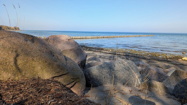 A rocky shoreline with large boulders, a wooden pier extending into calm water, and sparse vegetation.