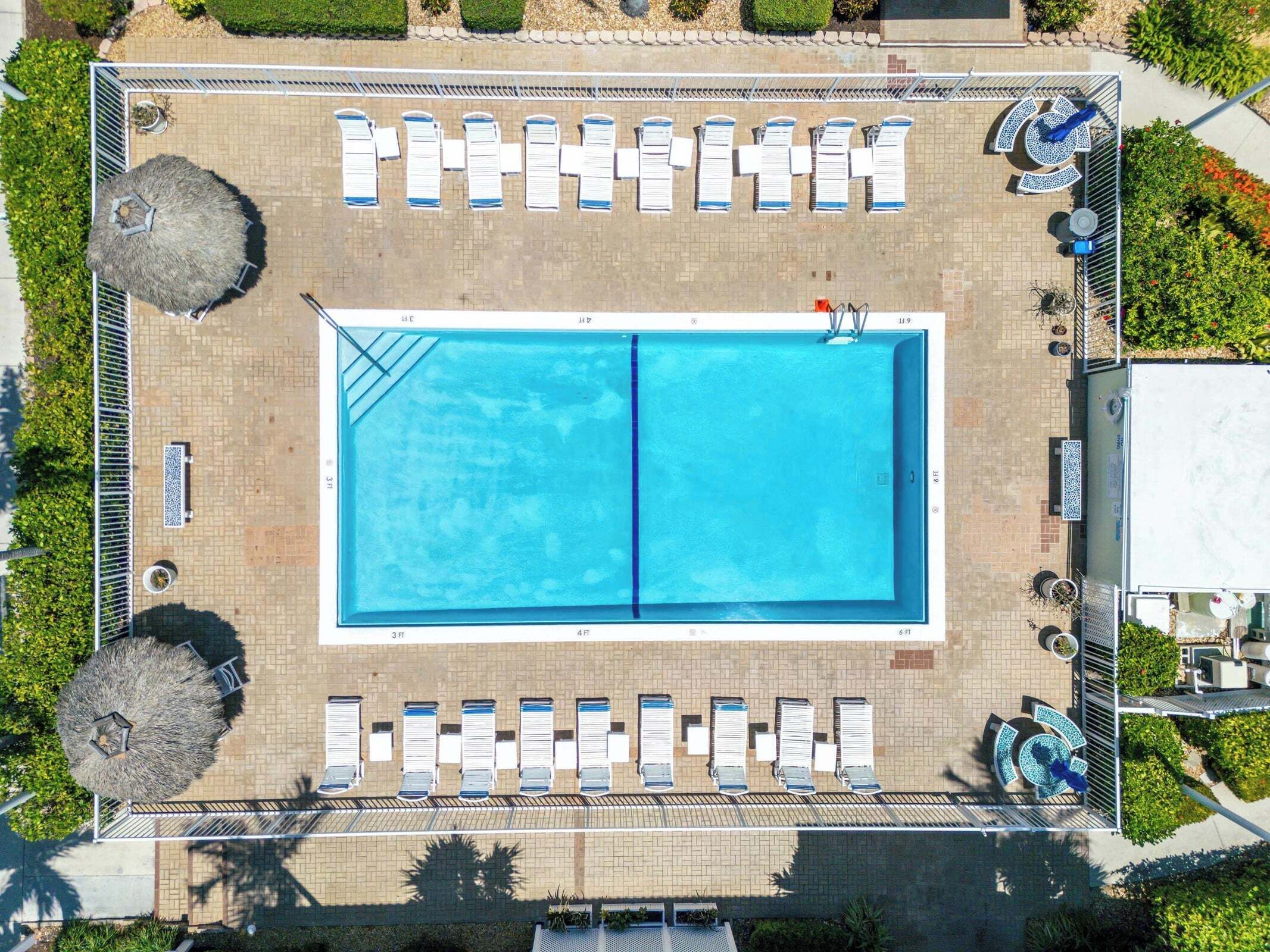 An aerial view of a rectangular swimming pool with lounge chairs arranged around it, surrounded by a paved walkway and greenery.
