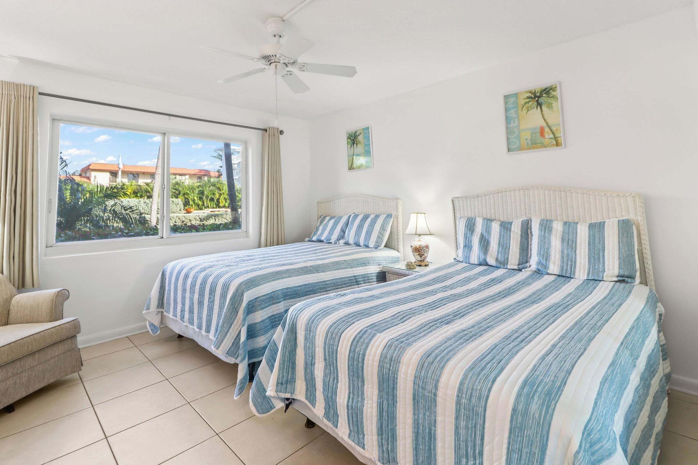 A room with two beds, a ceiling fan, a window with curtains, and a view of greenery outside.