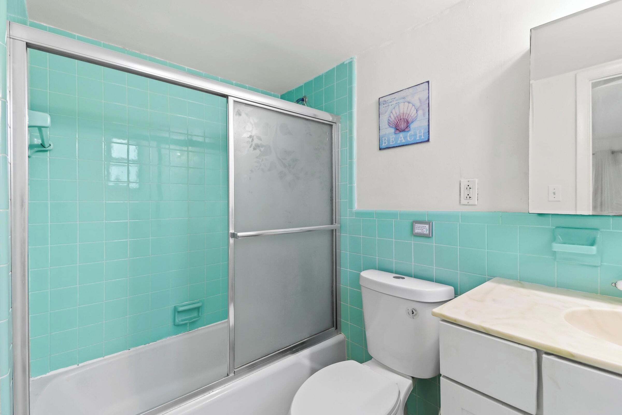 A bathroom with turquoise tiles, a white toilet, a shower with a frosted glass door, and a small framed picture on the wall.