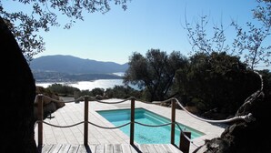 Eine Holzterrasse mit Pool, mit Blick auf eine Küstenlandschaft und Berge im Hintergrund.