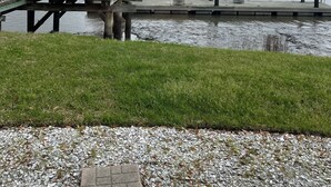 Een houten steiger met een overdekte zitplaats, uitkijkend over een wateroppervlak, omringd door grasvelden en een verre boomrij.