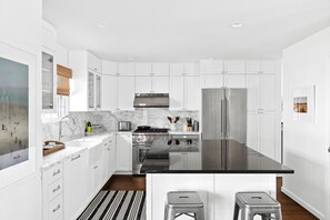 A modern kitchen with white cabinetry, stainless steel appliances, a marble countertop, and a black island with stools. - Classic 4-bedroom Home in East Hampton, Minutes from Town (East Hampton)
