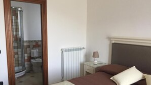 A bedroom with a bed, a nightstand, a lamp, a radiator, and a bathroom visible through an open door.
