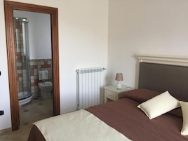 Un dormitorio con cama, mesita de noche, lámpara, radiador y baño visible a través de una puerta abierta.