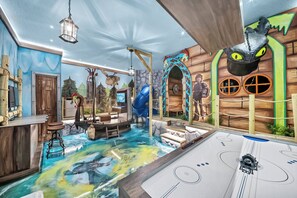 Ein Zimmer mit Spielbereich, einer Holztür, einem Schreibtisch und einem großen Fenster.