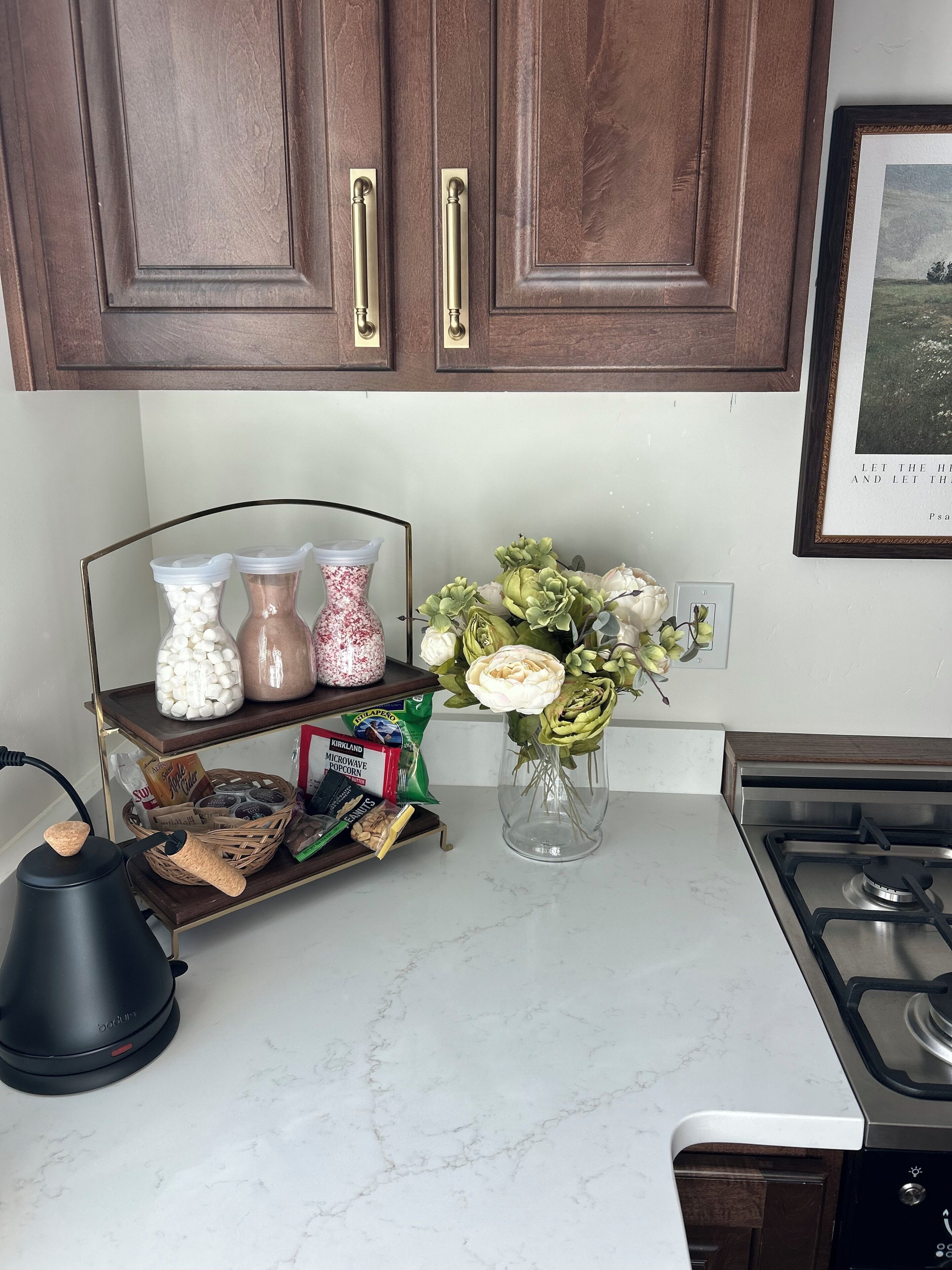 Une cuisine avec des meubles en bois, une cuisinière à gaz, une bouilloire, une étagère avec des bocaux et un vase rempli de fleurs.