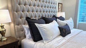 A bedroom with a tufted headboard, a bed with white and patterned bedding, a nightstand with a lamp, and a window with a view.