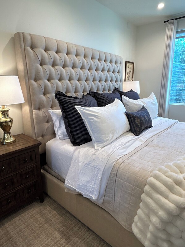A bedroom with a tufted headboard, a bed with white and patterned bedding, a nightstand with a lamp, and a window with a view.