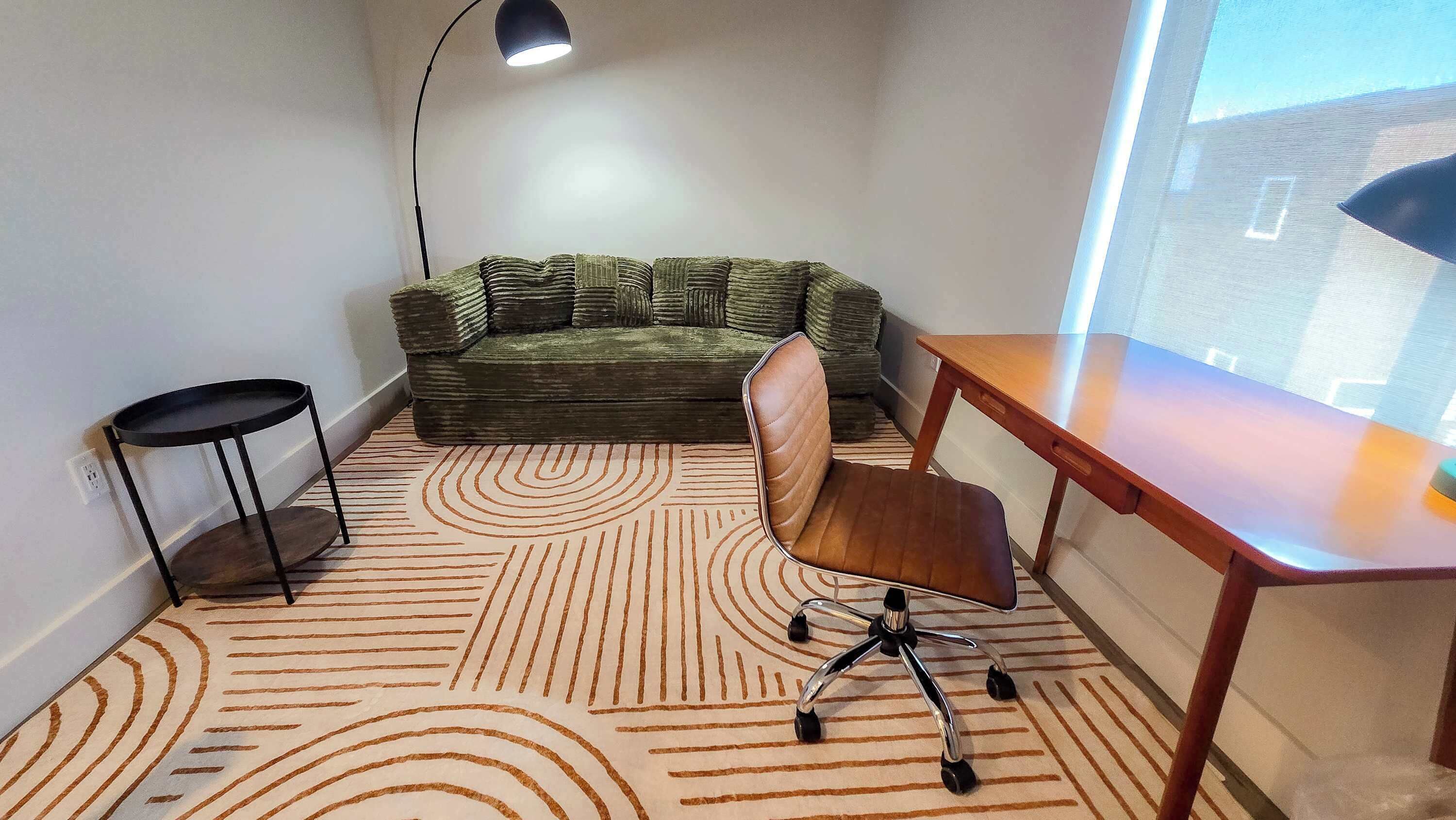 A small room with a green sofa, a wooden desk, a chair, a small table, and a patterned carpet.