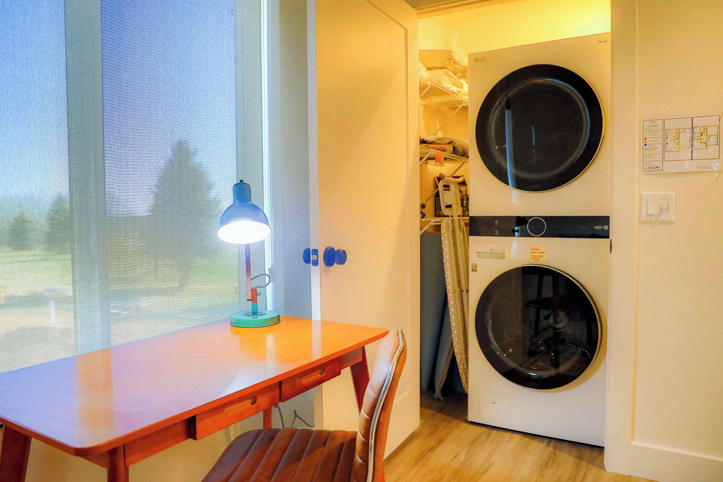 A room with a wooden table, a chair, a washing machine, and a window with a view of trees.