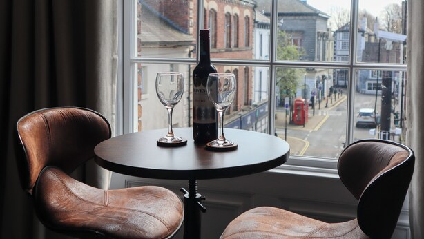 A table with a bottle and two glasses, two leather chairs, and a view of buildings through a window. - Knaresborough Retreat - Group Accommodation (Knaresborough)