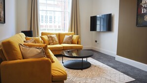 A living room with a yellow sofa, a round coffee table, a flat-screen TV mounted on the wall, and a patterned rug. - Knaresborough Retreat - Group Accommodation (Knaresborough)