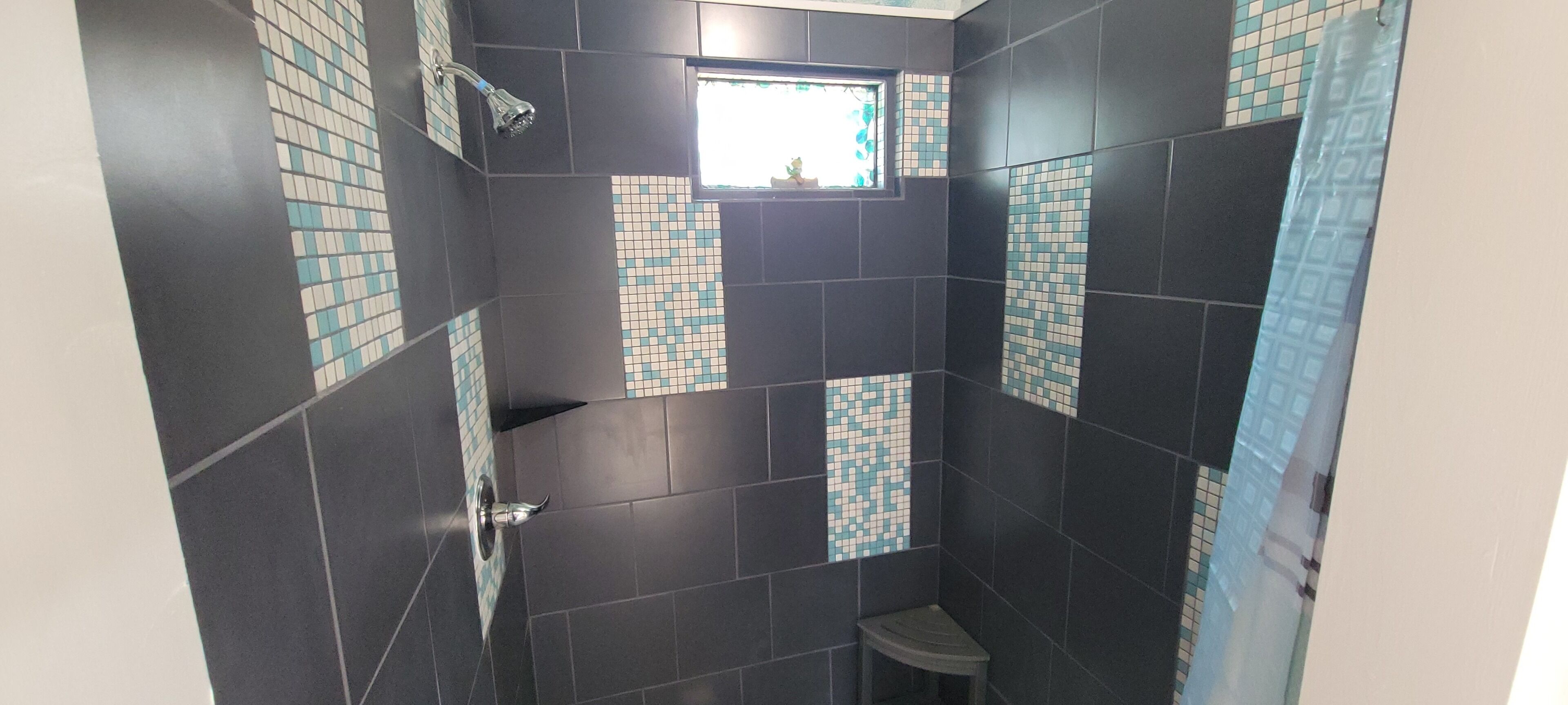 Une salle de bain avec des carreaux de couleur foncée, une douche, une fenêtre et un petit tabouret.
