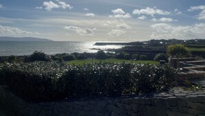 Paesaggio costiero con un muro in pietra, vegetazione e vista sul mare e colline lontane.