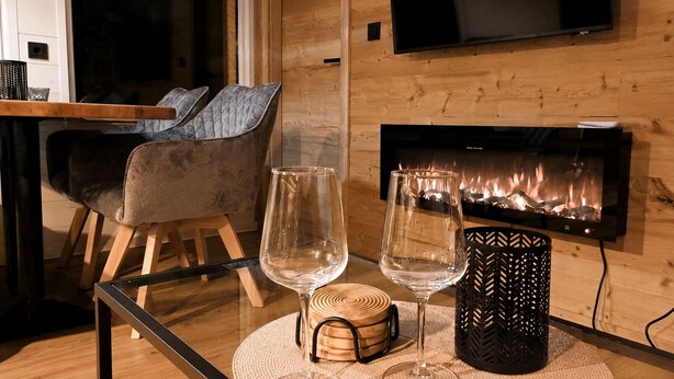 Un salon moderne avec une table basse en verre, deux verres à vin, un sous-verre en fibres tressées, un chandelier noir et une cheminée.