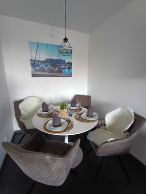 Un coin repas avec une table ronde dressée pour quatre personnes, entourée de chaises rembourrées, d’un luminaire suspendu et d’un tableau représentant un paysage urbain accroché au mur.