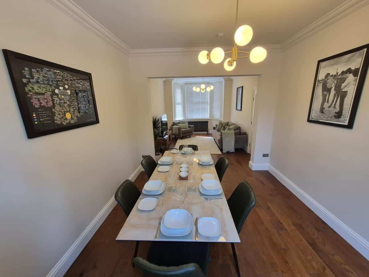 A dining room with a long table set for six, flanked by framed artwork, a chandelier, and a view into the adjacent living area.