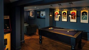 A billiards table with a pool cue, a dartboard, and framed album covers on the wall.