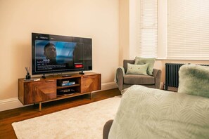 A modern living room with a flat-screen TV, a wooden entertainment unit, a grey sofa, and a white rug. - MCR Music Lover’s Dream 3BR House with Games Room! (Manchester)