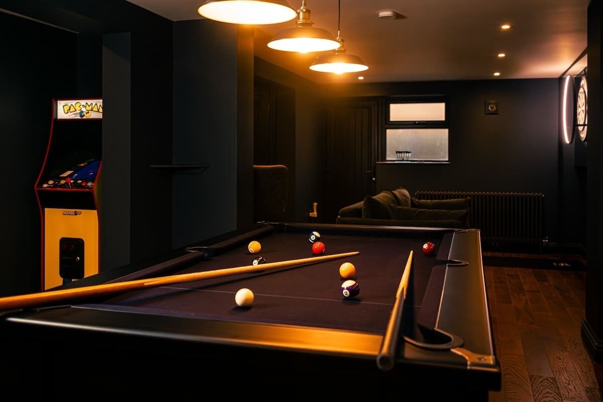 A pool table with balls, a pool cue, and a PAC-MAN arcade machine in a dimly lit room with a window and a radiator.